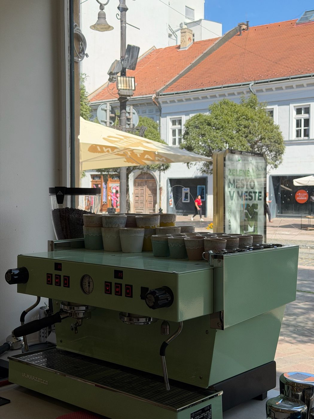 Pastelovo zelený kávovar La Marzocco pri okne s výhľadom na Hlavnú